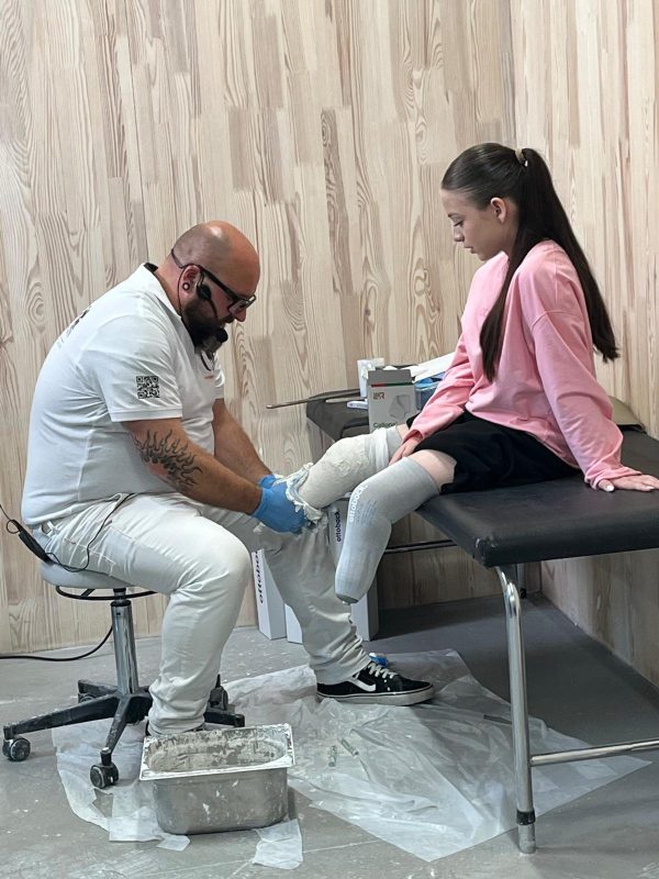 A specialist ist working on a orthesis of a girl in a pink sweater, who is sitting, looking down