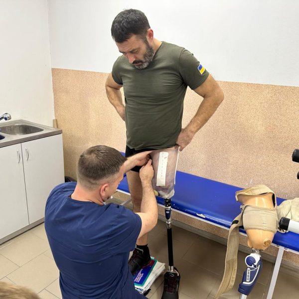 A person standing on one leg, getting a protesis on the other leg, is looking down to see the specialist helping him