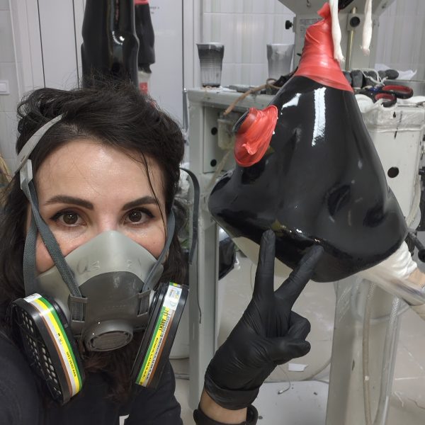 1000016429 A woman wearing a respirator mask and black gloves poses for a selfie in a workshop, pointing at a black, cone-shaped object covered in red plastic wrap.
The woman, positioned in the foreground on the left side of the frame, has dark hair and wears a grey respirator mask. The mask covers her nose and mouth, and it has prominent filters attached to the sides. She wears black, form-fitting gloves, and her left hand is raised, with two fingers extended in a peace sign, pointing towards the object. A black bracelet is visible on her right wrist. She is wearing a black t-shirt.
The main subject of the image, besides the woman, is the black, glossy, cone-shaped object. It appears to be made of a hard material and is partially covered with bright red plastic wrap, particularly at its upper section and a smaller protrusion near what might be considered its base. Two white ropes hang from the top of the object. The object is suspended or mounted on some sort of equipment, visible behind it.
The setting appears to be a workshop. The background consists of white tiled walls and some industrial-looking equipment. To the left, a white door with vertical lines is visible. On a surface behind the object, there are some disposable cups, one of which is grey and appears to have paint or residue inside. Other tools or materials are scattered around, suggesting work in progress. The overall atmosphere is practical and focused on the task at hand, with the woman’s direct gaze and gesture adding a touch of personal engagement to the scene.