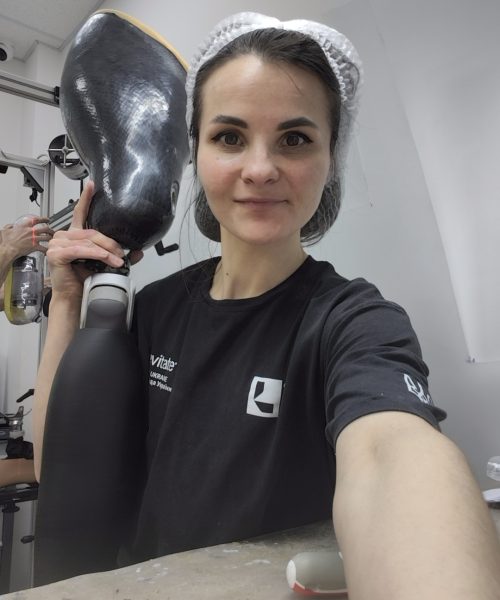 Olena Fomenko The image shows a woman in a workshop, holding a prosthetic limb. She is wearing a black t-shirt with a white logo and a white hairnet. The prosthetic limb is dark gray and appears to be made of a composite material. It consists of a socket and a lower shaft. The woman is smiling and looking at the camera. The background of the image is a workshop environment. To the left, there are some machines and equipment, including what looks like a stand with some cylindrical objects. A partial view of another person's arm and hand can be seen on the far left, possibly working on something. The wall behind the woman is white and there is a ceiling with fluorescent lighting. In the foreground, on a textured workbench, there are several objects. These include a metallic fitting on the left and some white and gray components of what might be other prosthetics or related equipment on the right. The workbench itself is cluttered with various materials, possibly remnants of the prosthetic fabrication process, such as small yellow and white specks. The composition is a close-up shot, with the woman occupying a significant portion of the frame. She is positioned slightly off-center to the right, and the prosthetic limb is held up in the left part of the frame, creating a balanced diagonal line. The perspective is from a slightly low angle, looking up at the woman. The focus is sharp on the woman and the prosthetic limb she is holding, with the background slightly blurred. Subtle details include the texture of the prosthetic limb's socket, which appears to be woven carbon fiber, and the details on the various fittings on the workbench.